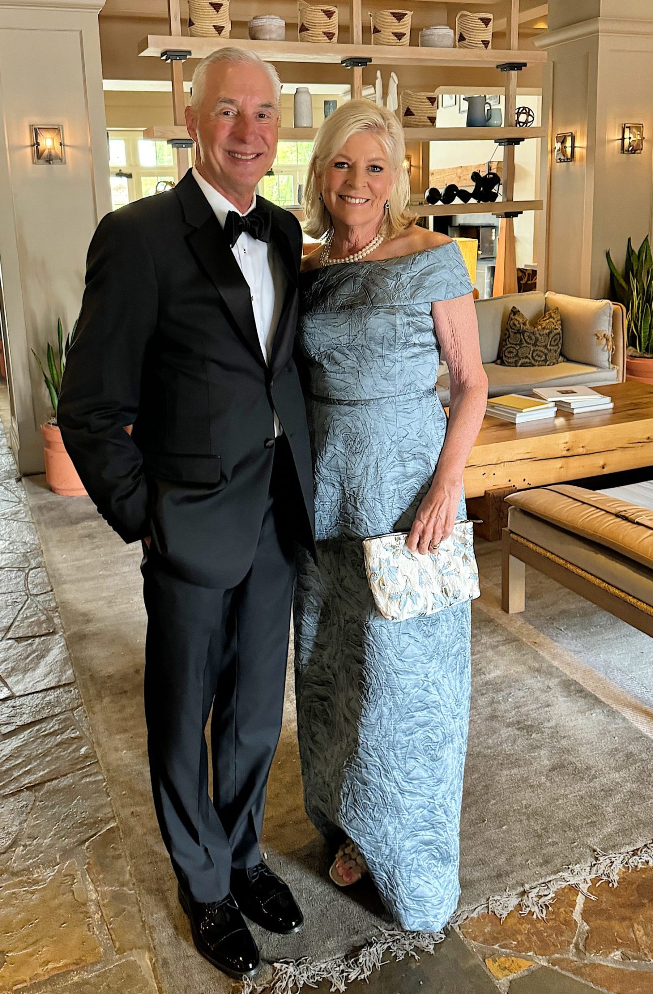 A man in a black tuxedo and woman in a blue gown stand together in a living room.