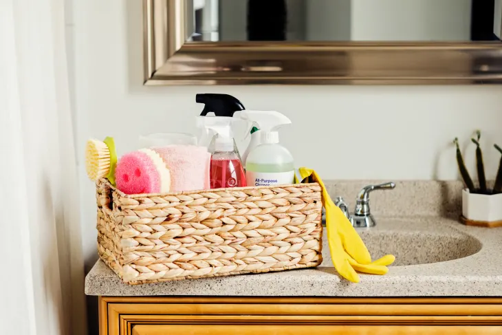 A woven basket with cleaning supplies, including spray bottles, sponges, and a brush, sits on a bathroom counter next to a silver faucet.