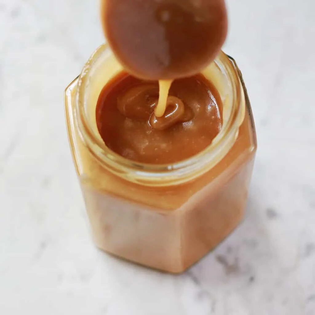 A jar filled with caramel sauce, with a spoon hovering above, dripping caramel. The jar is on a light marble surface.