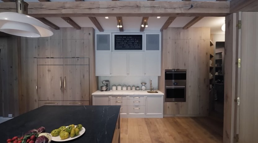 A modern kitchen with wooden cabinets, a large island counter, built-in stainless steel appliances, and assorted snacks on the counter. Exposed ceiling beams highlight the rustic design.