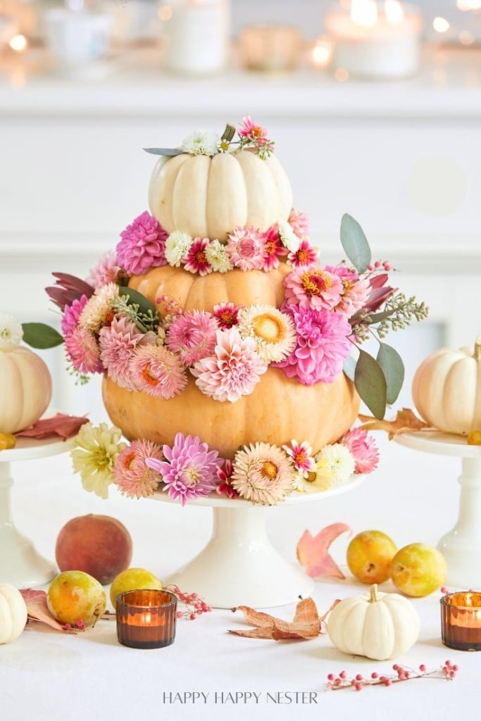 A tiered pumpkin and flower centerpiece on a cake stand, surrounded by small pumpkins, apples, and candles on a table.