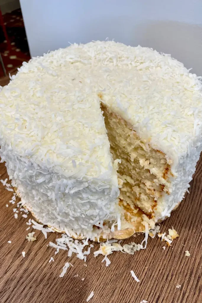 A round cake covered in white frosting and shredded coconut, with a large slice taken out, on a wooden surface.