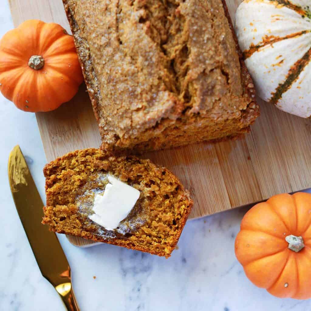 A loaf of pumpkin bread with a buttered slice on a wooden board, surrounded by small pumpkins. A gold knife is placed nearby.