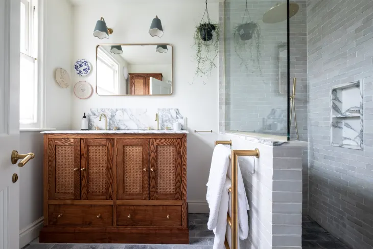 Bathroom with a wooden vanity, marble countertop, rectangular mirror, wall-mounted lights, a glass shower enclosure, and hanging plant decor.