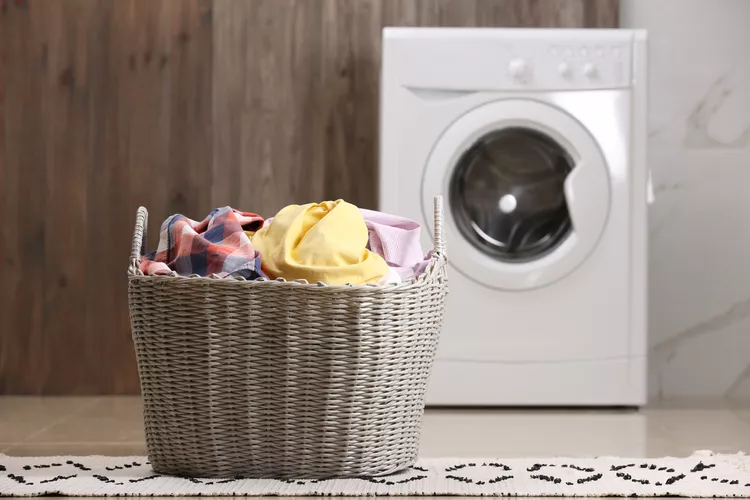 A wicker laundry basket filled with colorful clothes sits on the floor in front of a washing machine.