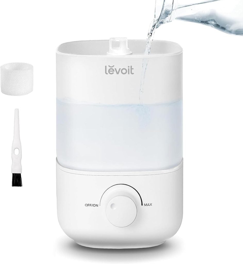 White Levoit humidifier with water being poured in, resembling a classic nutcracker Christmas charm. It features a dial for settings and includes a small brush and filter beside it.