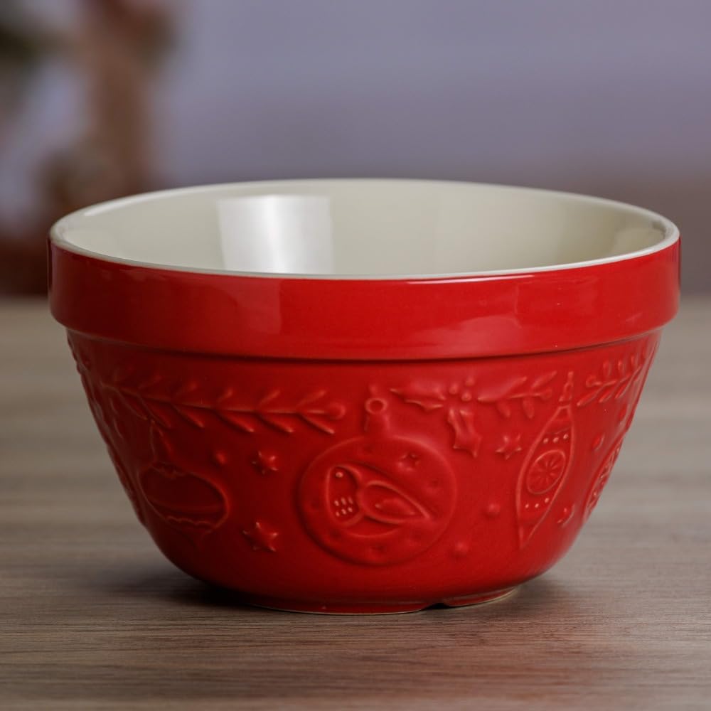 A red ceramic bowl with embossed holiday designs sits on a wooden surface, its cream-colored interior perfect for mixing batter for Christmas tree cupcakes.