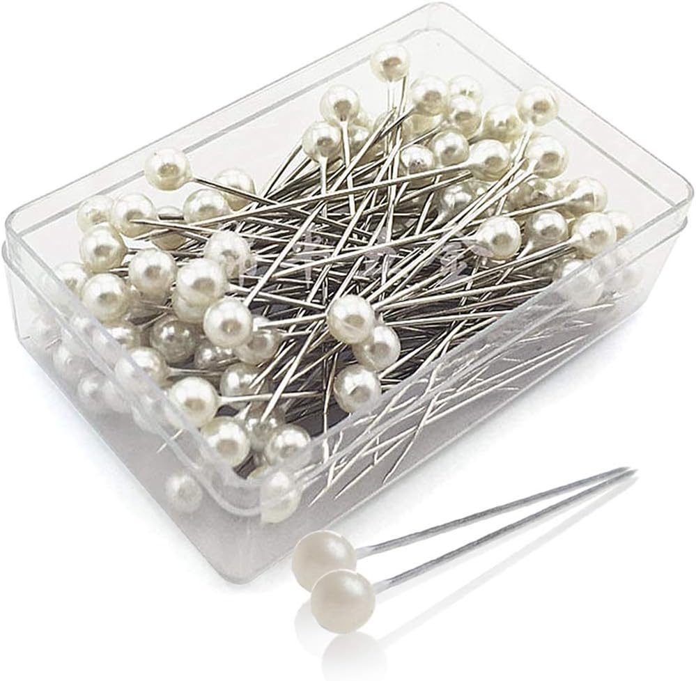 A clear plastic box filled with silver straight pins, each topped with a small white bead, perfect for securing your festive Christmas tree skirt. Two loose pins are placed beside the box.