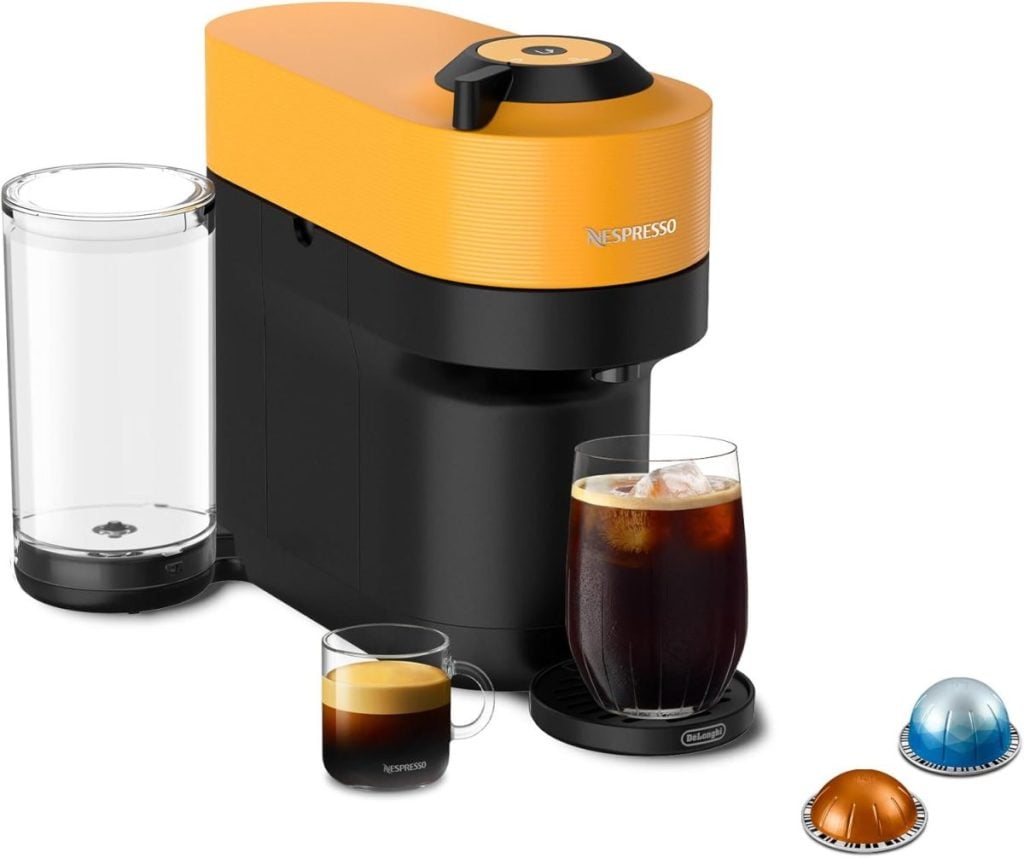 An orange and black Nespresso machine, reminiscent of a nutcracker in its precision, sits with a water reservoir, a small espresso cup, a festive glass of iced coffee, and three coffee pods ready to bring holiday cheer.