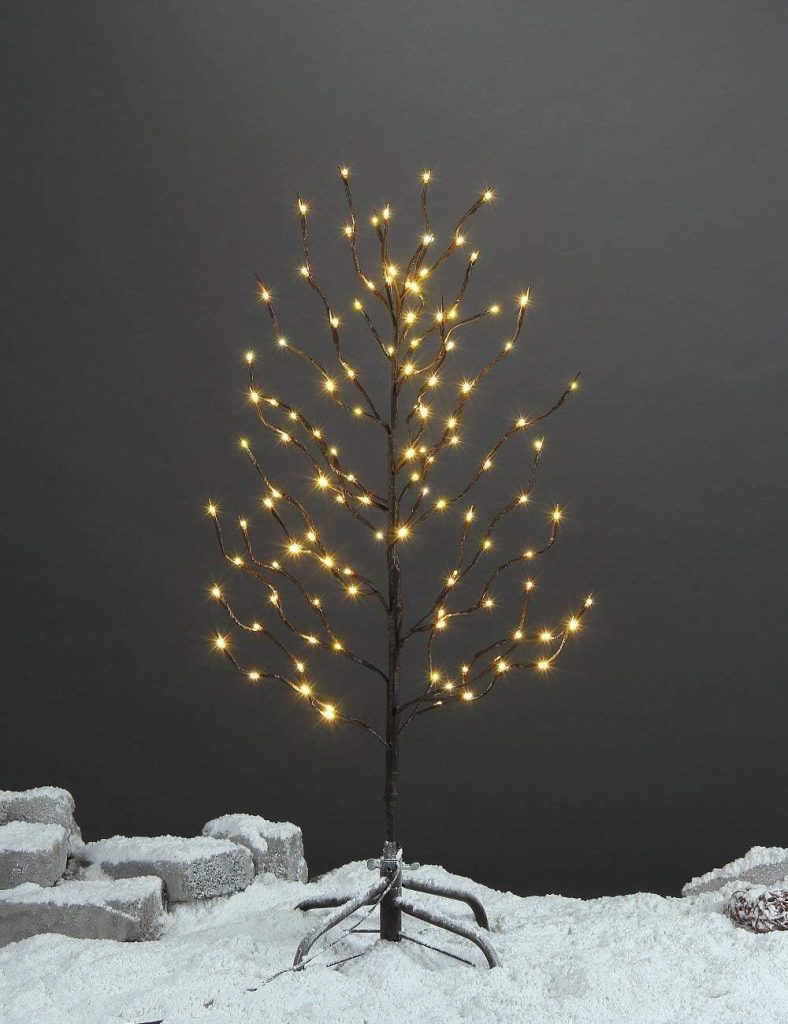 A leafless artificial tree with bare branches is decorated with small warm white lights, standing on a snowy surface with stone blocks in the background—perfect for creating a festive atmosphere this Black Friday.