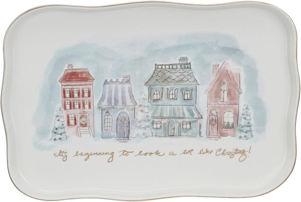 A rectangular plate features a painted row of four winter houses with trees, including touches of pink Christmas decor, and the phrase "It's beginning to look a lot like Christmas!" written in cursive at the bottom.