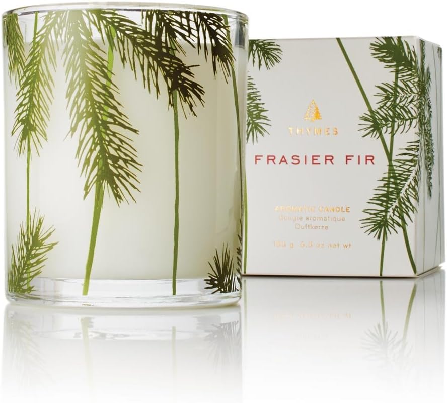 A candle in a glass holder with green pine branch designs, next to a box labeled "Frasier Fir" by Thymes.