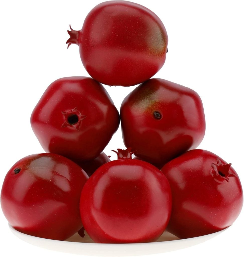 Six shiny red pomegranates, adorned with a festive Christmas ribbon, are stacked in a pyramid formation on a white plate.