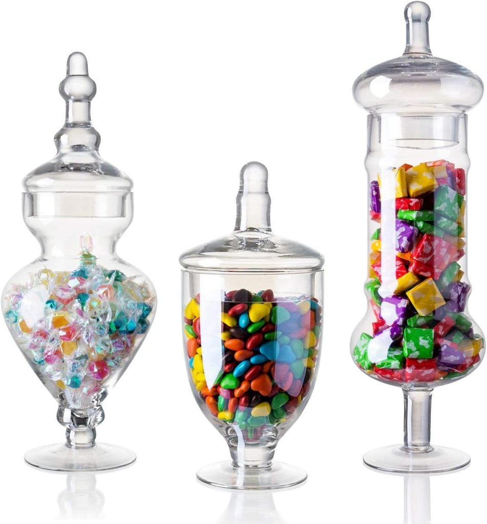 Three glass candy jars filled with assorted candies; one with wrapped hard candies, another with colorful candy-coated chocolates, and the third with wrapped square sweets.