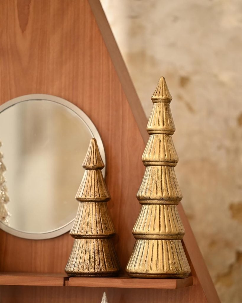 Two gold, cone-shaped decorative objects resembling stylized trees, adorned with subtle Christmas colors, are placed on a wooden shelf near a round mirror.