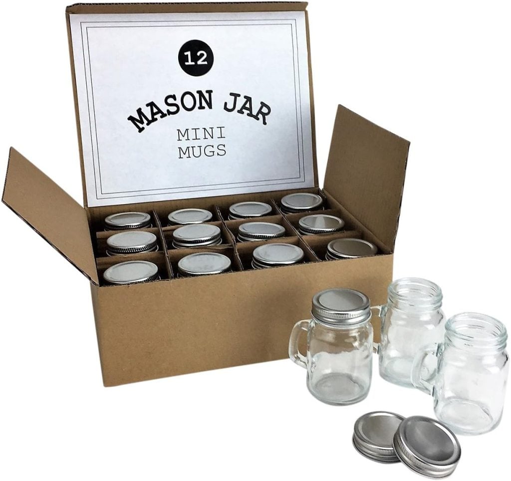 A cardboard box containing 12 mini Mason jar mugs with silver lids, perfect for festive gatherings. Three jars and three lids, adorned with a touch of Christmas ribbon, are displayed outside the box.