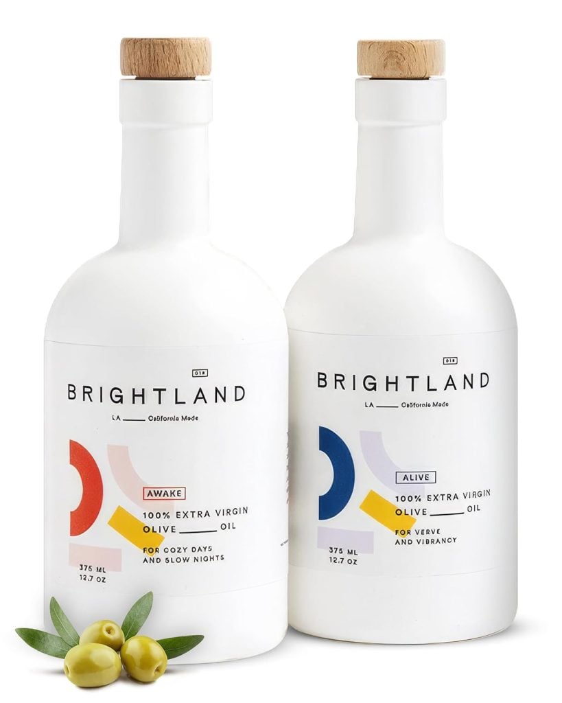 Two bottles of Brightland extra virgin olive oil, labeled "Awake" and "Alive," with a few olives and leaves beside them.