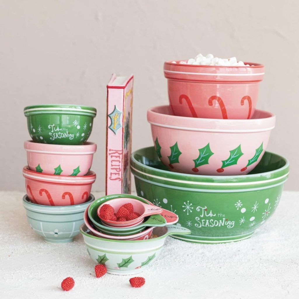 A set of festive mixing bowls in pink and green with holiday designs, perfect for preparing holiday desserts, includes a recipe book and small candies tucked inside some bowls, all arranged on a white surface.