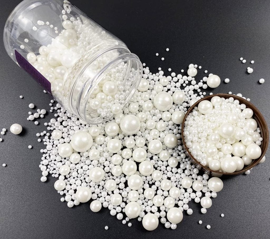 A plastic jar is tipped over, spilling various sized white pearls onto a black surface, reminiscent of sugar decorations on Christmas tree cupcakes. A small bowl is filled with similar pearls.