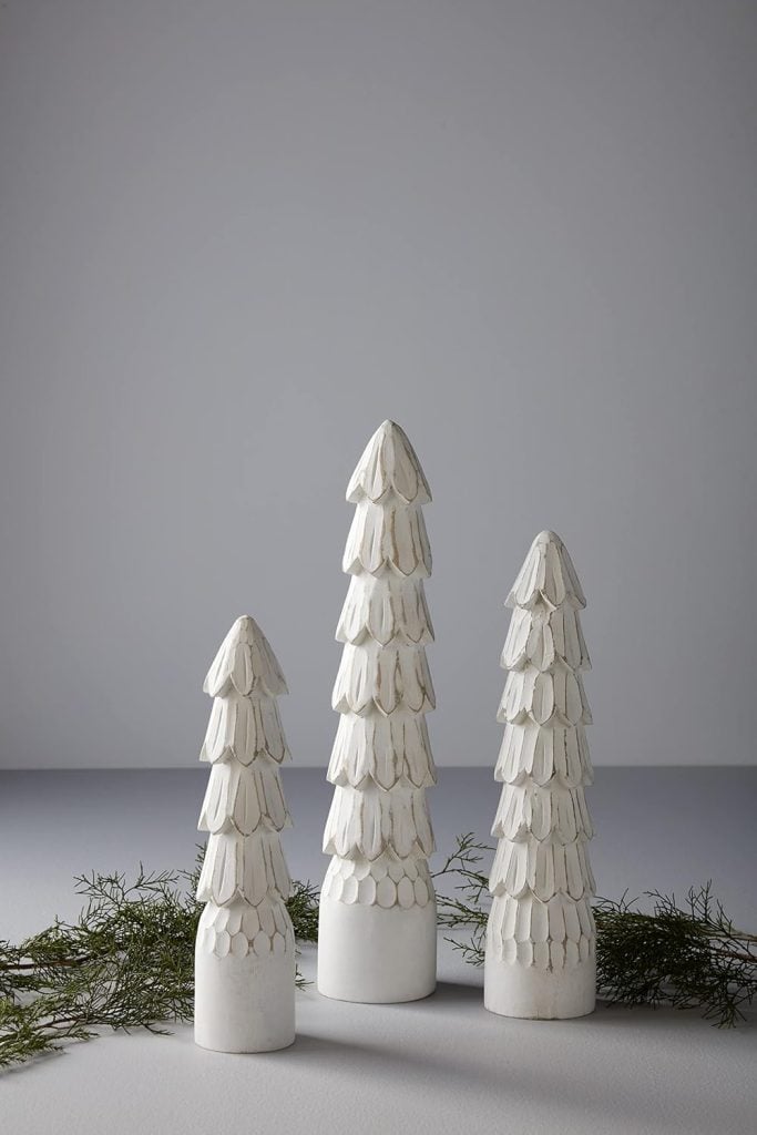 Three white, cone-shaped sculptures resembling trees are arranged in a row on a gray surface, with green foliage and button crafts thoughtfully scattered around.