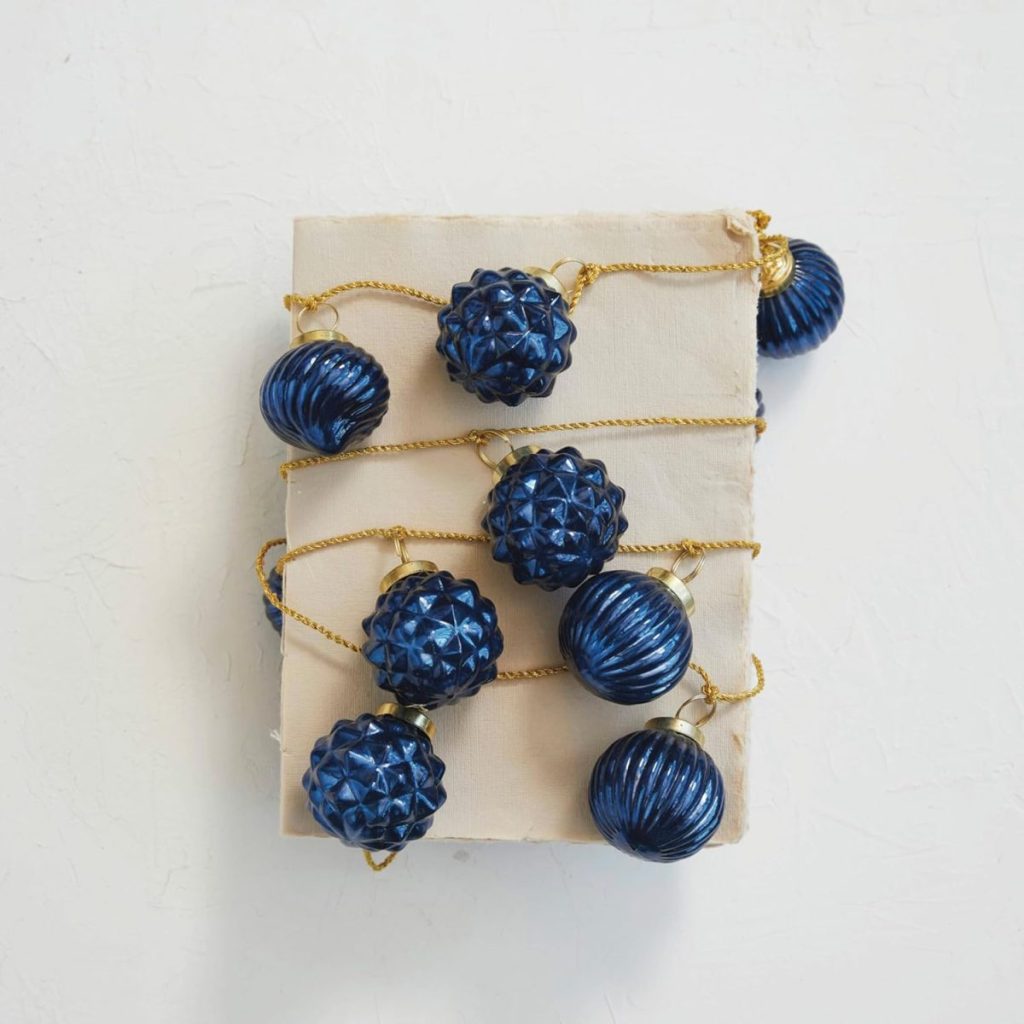 A string of blue, textured baubles with gold caps and cord evokes a Beach Christmas vibe as they rest atop a wrapped, rectangular package against a white background.