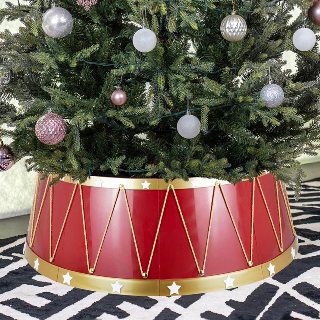 A Christmas tree with a red drum-shaped tree collar sits gracefully on a patterned rug, adorned with silver and pink ornaments, alongside a delicate Christmas tree skirt for added charm.