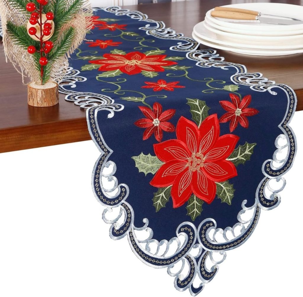 A festive table runner adorned with Christmas colors, featuring red poinsettia patterns and ornate lace edges, graces a wooden table with stacked white plates on top.