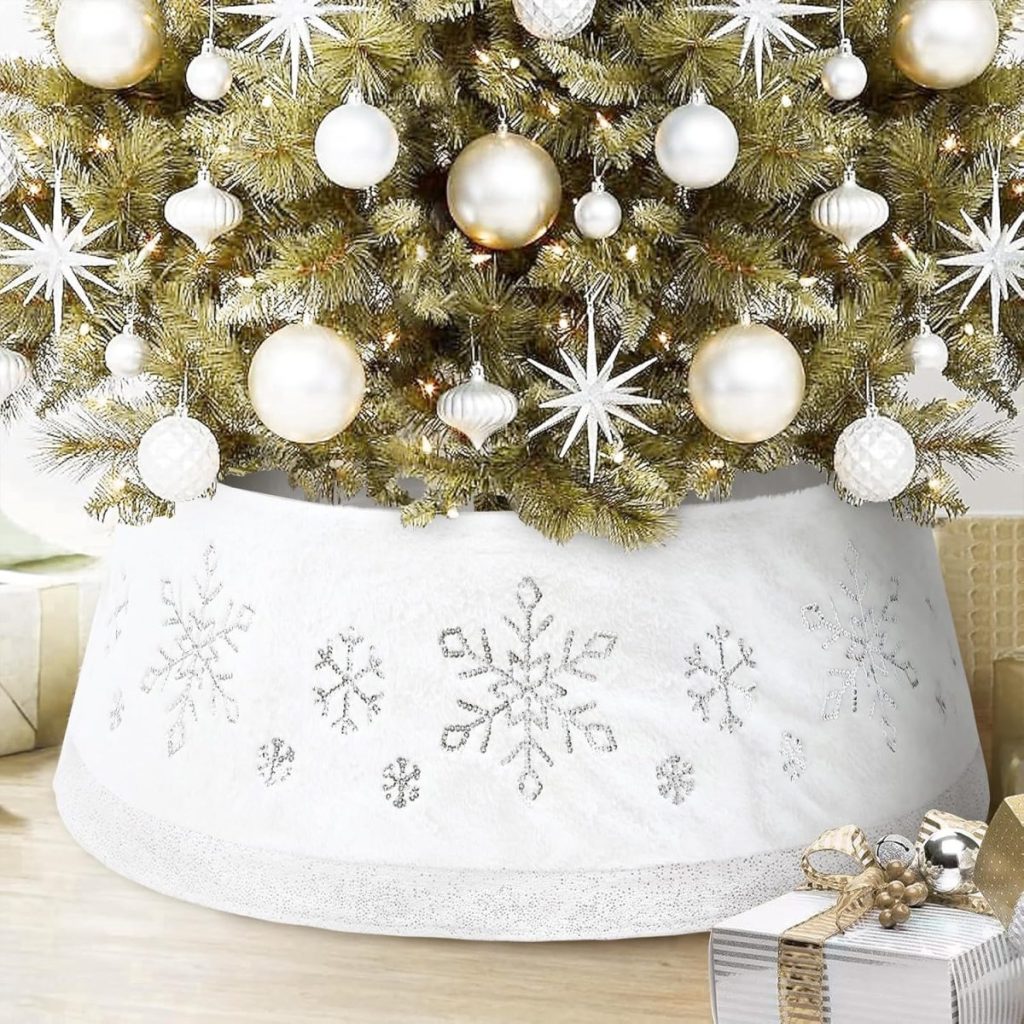 A decorated Christmas tree with white and gold ornaments, featuring a christmas tree skirt adorned with silver snowflake patterns, stands next to wrapped gifts.