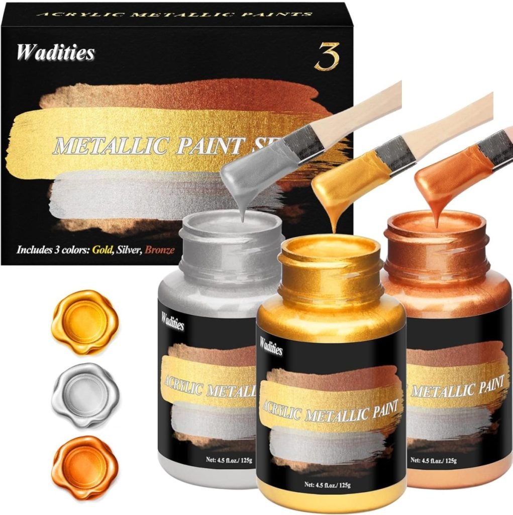 Acrylic metallic paint set with three bottles in gold, silver, and bronze, each paired with a brush. The packaging features color samples reminiscent of oyster shells and labels the set "Wadities Metallic Paint Set.