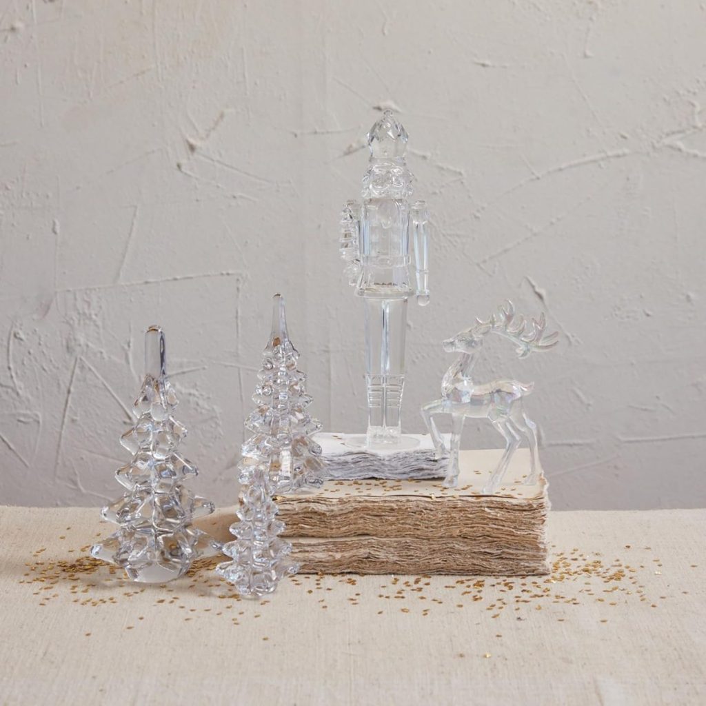 Four clear glass figurines—a nutcracker, a reindeer, and two trees—are arranged on a stack of textured paper, evoking the crisp clarity of a vodka cranberry against a plain, light-colored wall.
