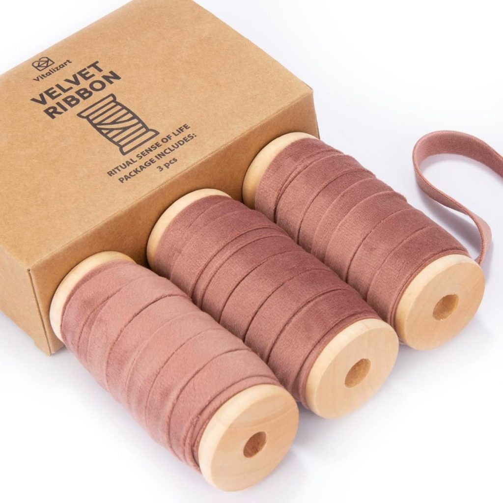 Three spools of dusty pink velvet ribbon in wooden reels are partially pulled from a cardboard box labeled "Vividchart Velvet Ribbon," perfect for those seeking the finest material on how to tie a bow with elegance.