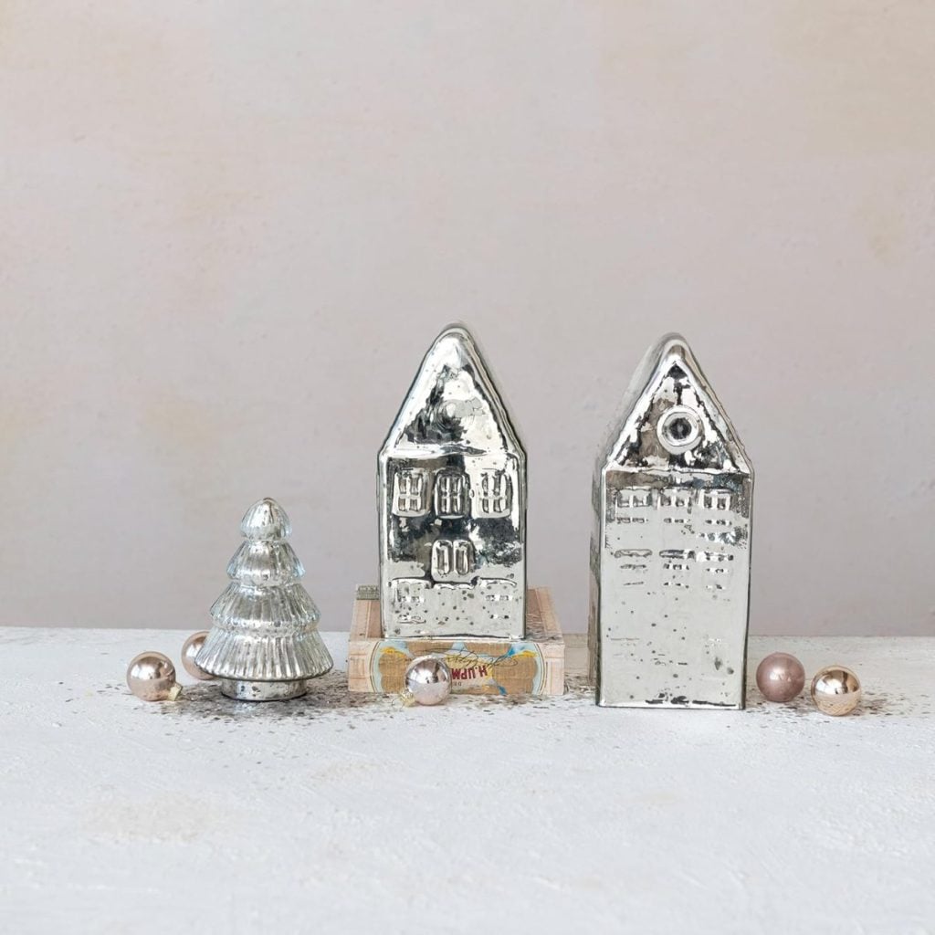 Set a Christmas table with two metallic house-shaped ornaments, a small glass Christmas tree, and several round decorative beads arranged on a white textured surface against a plain light background.