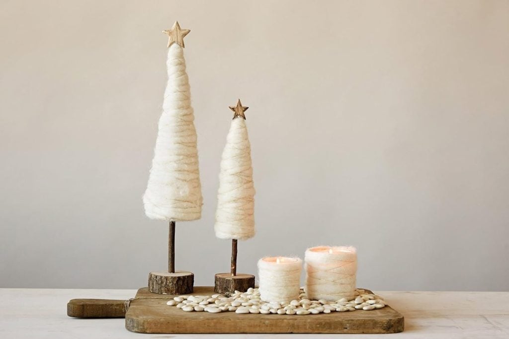 Two wool-wrapped conical trees in festive Christmas colors, topped with stars, stand on a wooden board beside two candles surrounded by white pebbles.