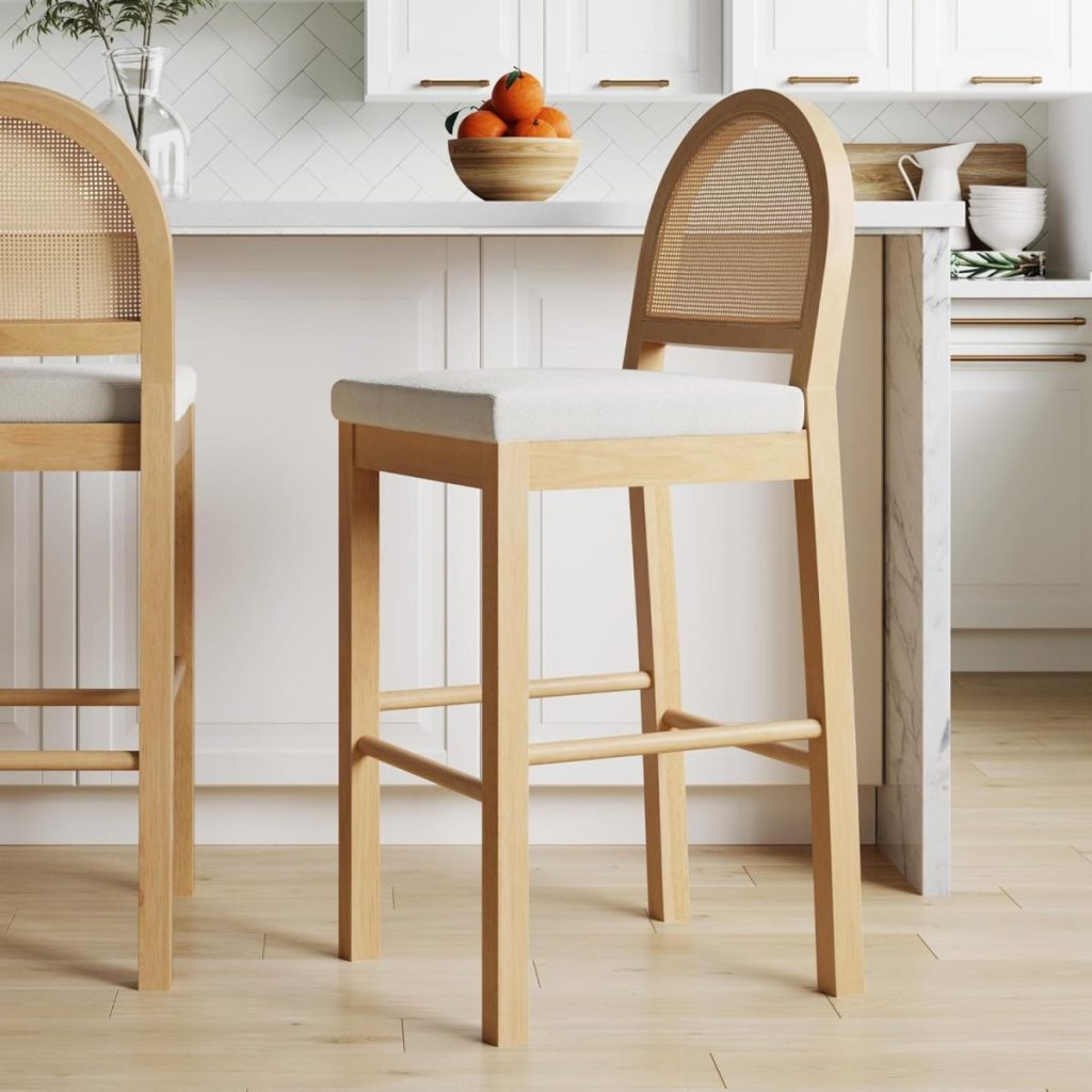 A wooden bar stool with a cushioned seat is positioned in a modern kitchen featuring sleek white cabinets and a marble countertop, ready for the Black Friday buzz. A bowl of oranges adds a pop of color on the counter.