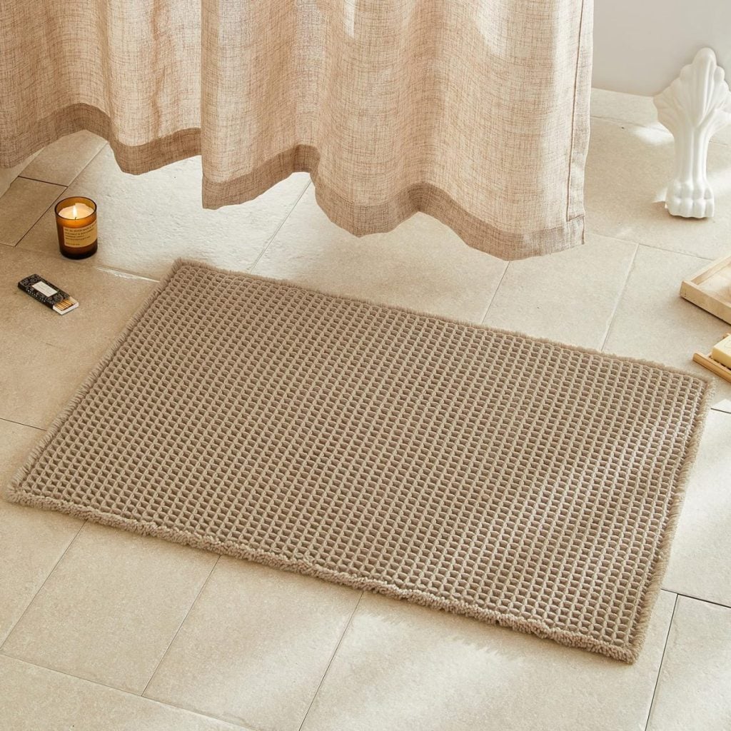 A beige waffle-textured bath mat is placed on a tiled bathroom floor near a light candle, a lighter, a statue, and a beige curtain—perfect for cozy Black Friday relaxation.