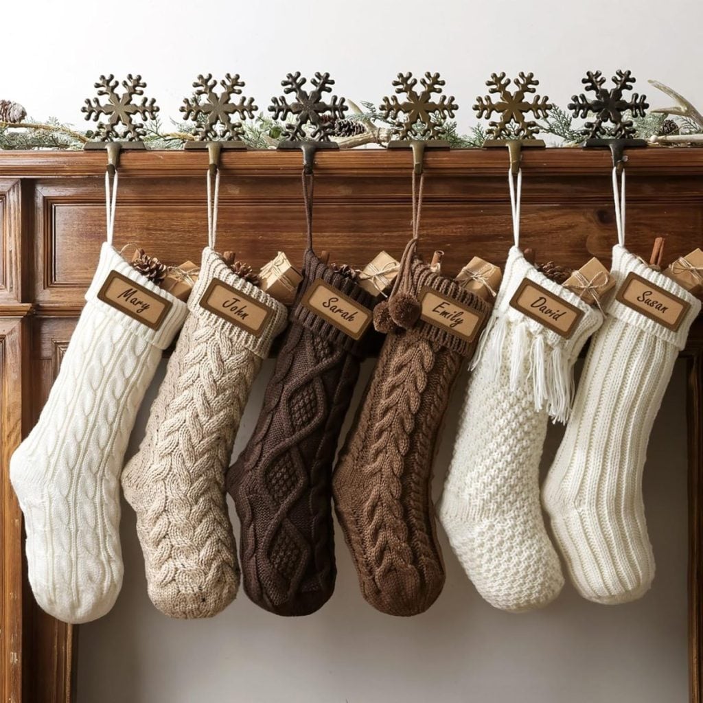 Six knit Christmas stockings in neutral colors hang from a mantel, each labeled with a wooden name tag and filled with small wrapped gifts, ready to delight everyone on Christmas morning.