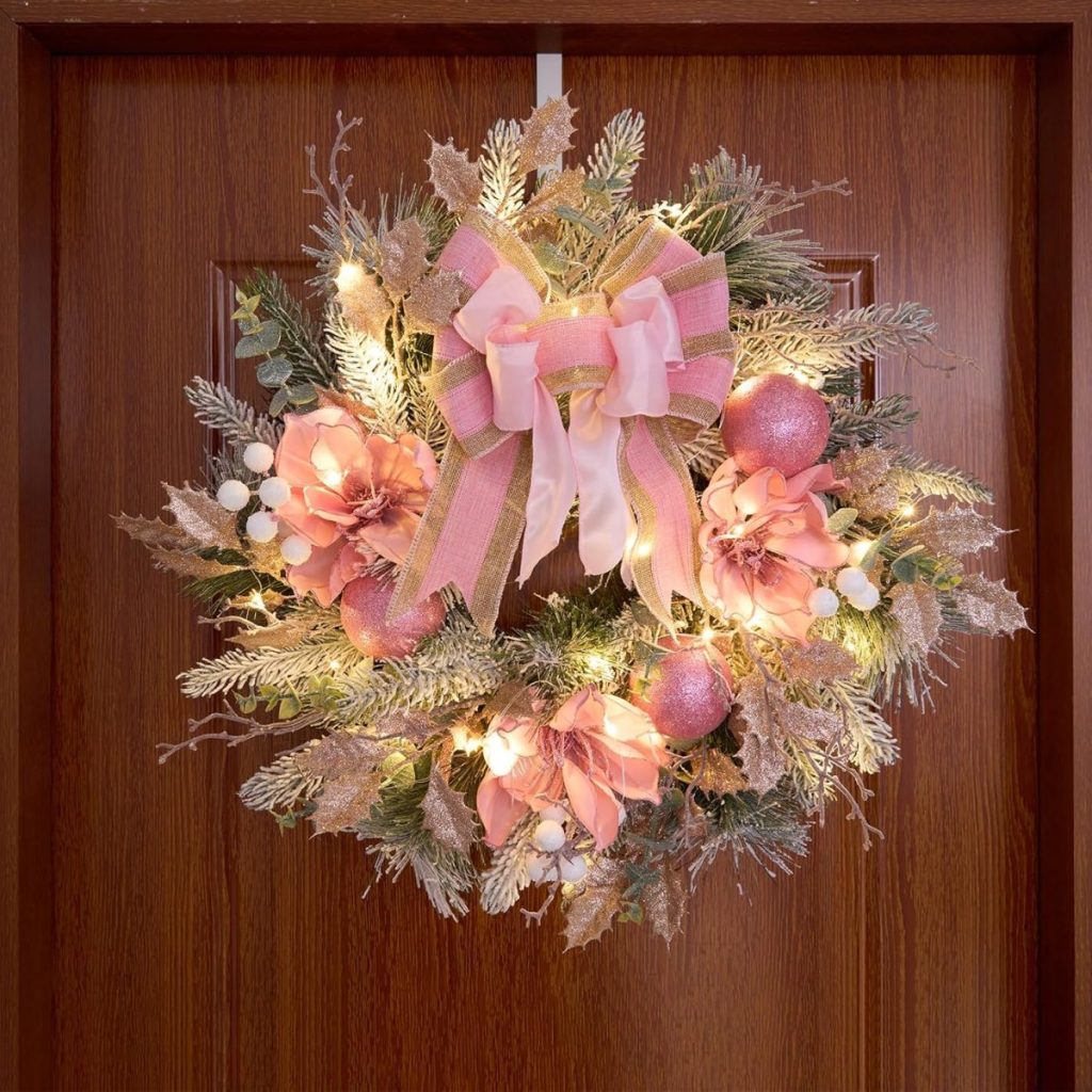 A festive Christmas wreath adorned with pink flowers, a large pink bow, and twinkling lights is elegantly hanging on a wooden door.