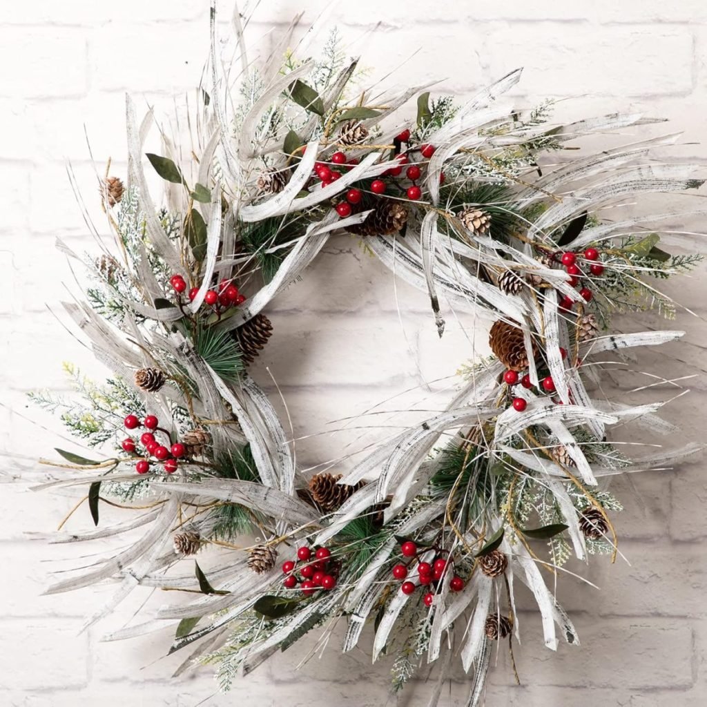 A Christmas wreath adorned with red berries, pinecones, and silvery foliage graces a white brick wall.