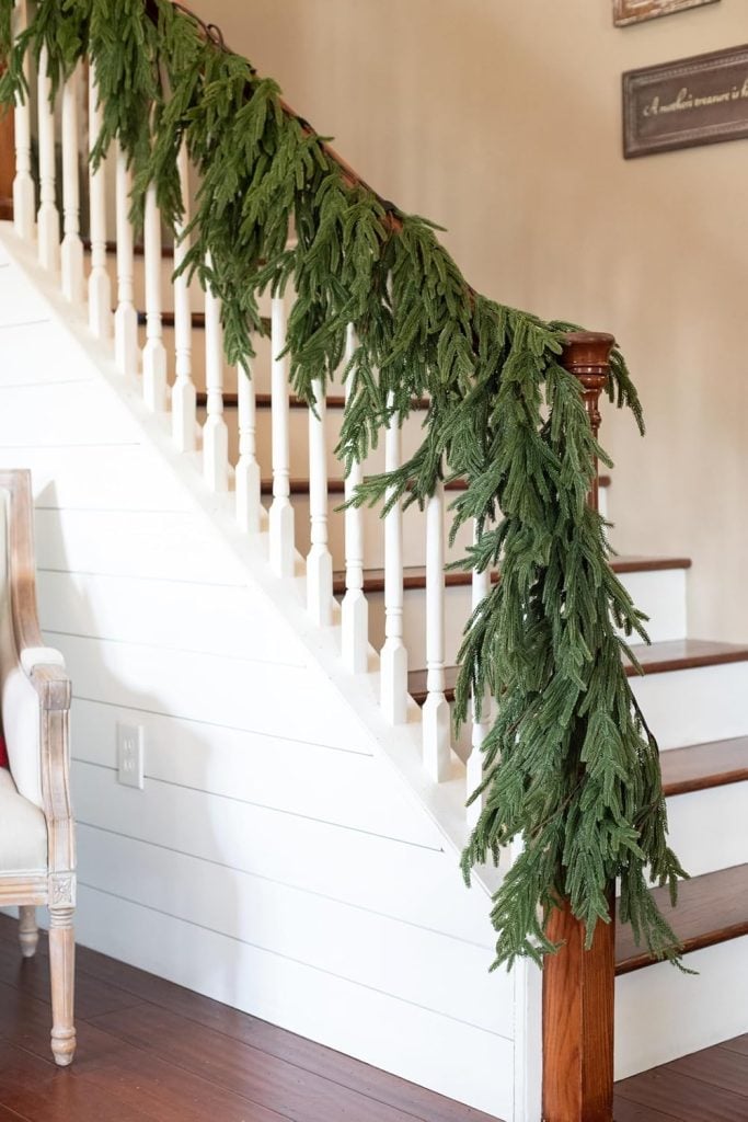 A staircase with white railings is adorned in Christmas colors, featuring green garland draped along the banister. An armchair with a wooden frame sits nearby, completing the festive atmosphere.