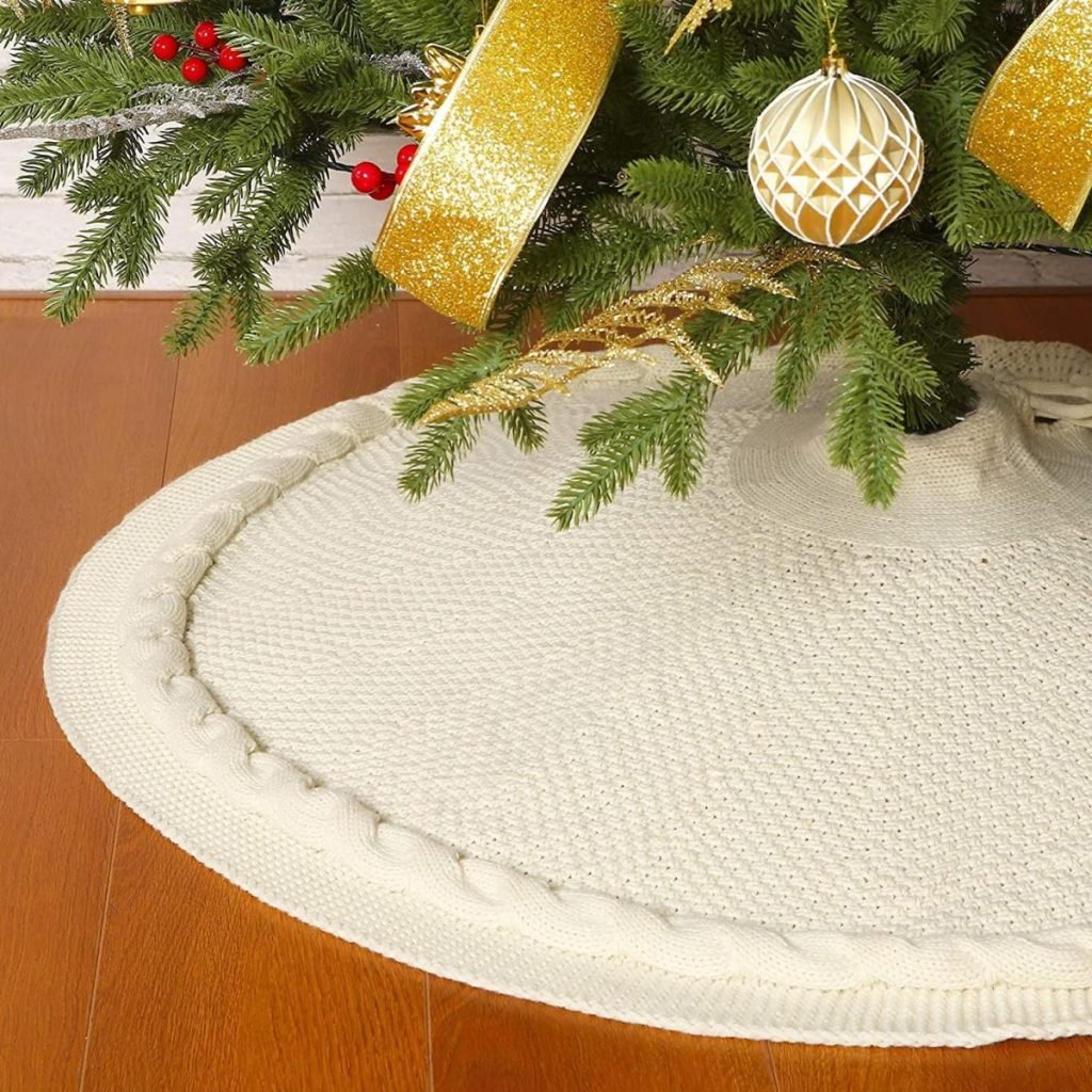 A Christmas tree on a white knit tree skirt glistens with gold ribbons and an elegant ornament, capturing the essence of Christmas colors amidst pine cones on a wooden floor.