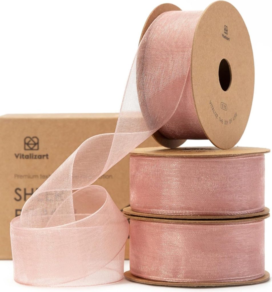 Three rolls of pink sheer ribbon are stacked, with one gracefully unrolled, in front of a cardboard box featuring the Vitalizart logo—a perfect setup to explore how to tie a bow with elegance and flair.