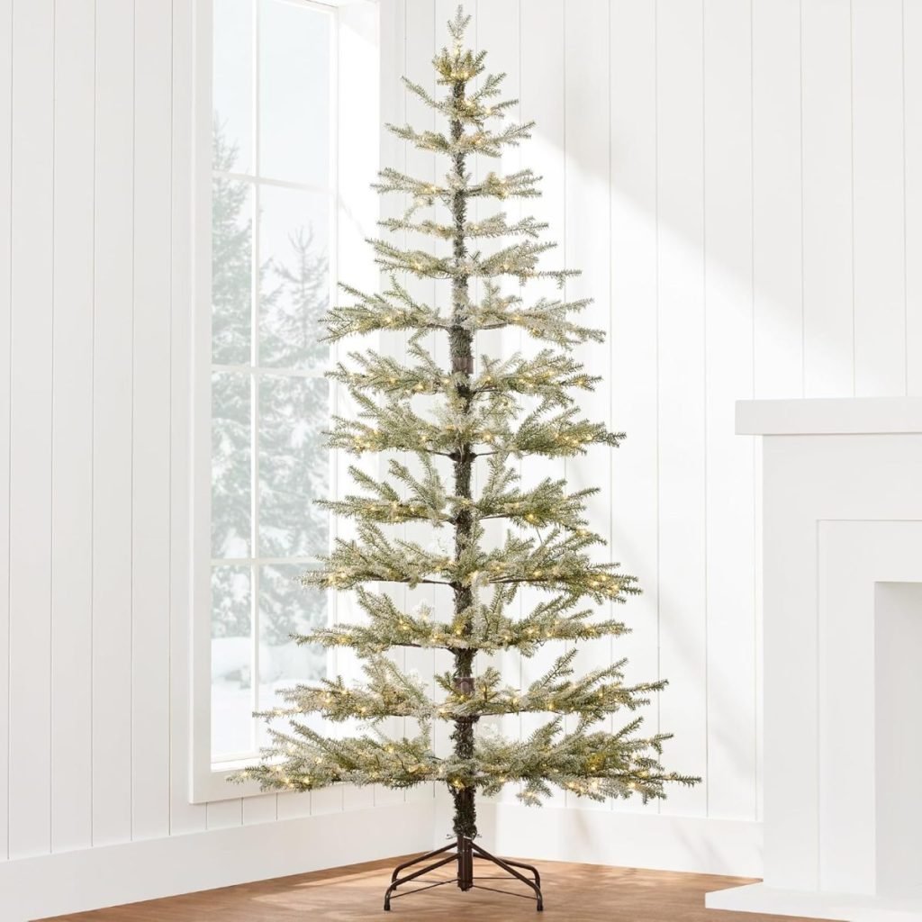 A slim, artificial, pre-lit Christmas tree stands in a bright room with white walls and a large window.