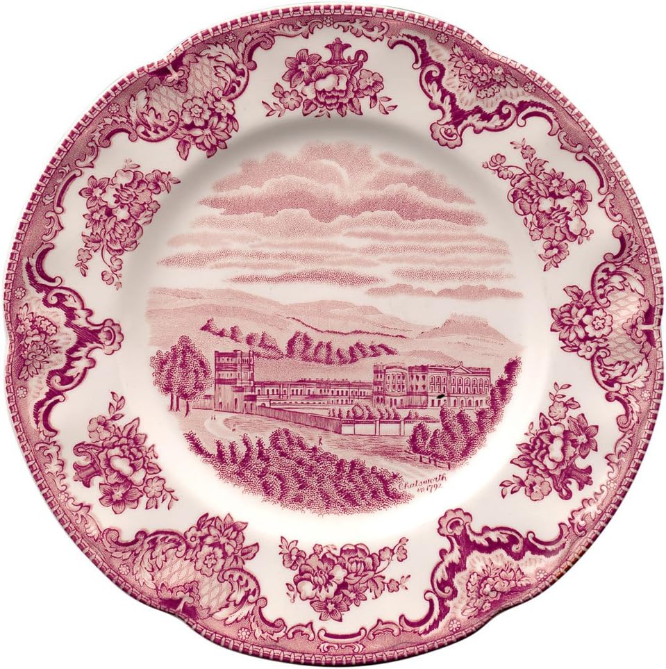 Ornate ceramic plate with a pink floral border in soft Christmas colors and a central illustration of buildings and a garden, set against a backdrop of hills and clouds.