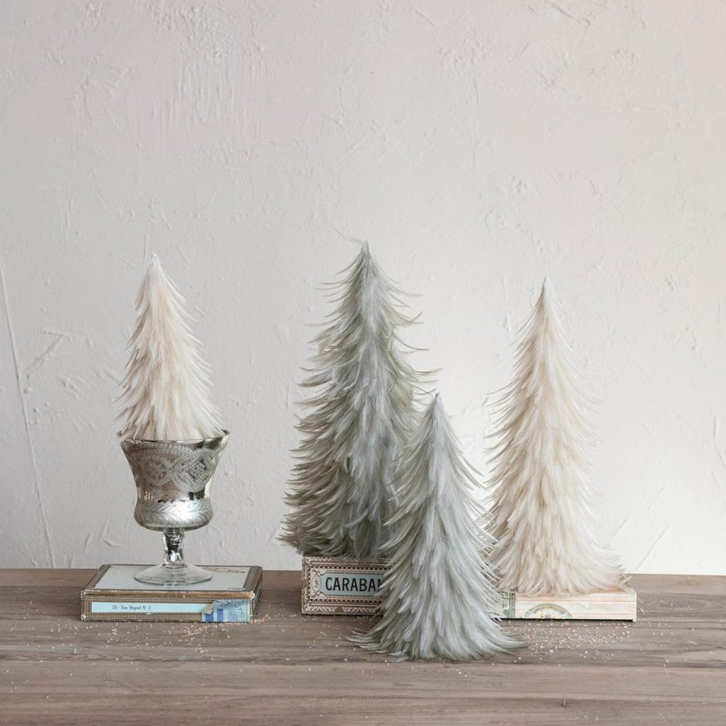 Four feathered decorative trees in white and gray stand on a wooden surface, resembling elegant Christmas tree cupcakes, with two positioned on stacked books and one in a silver cup.