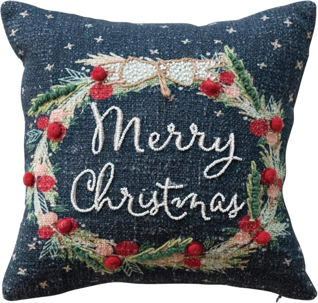 Decorative pillow featuring embroidered text "Merry Christmas" encircled by a wreath of red berries and lush greenery, capturing classic Christmas colors against a dark backdrop.