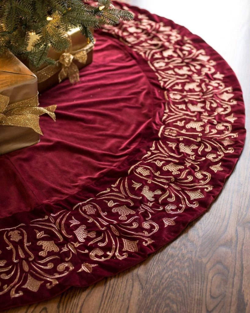 A red velvet christmas tree skirt, adorned with golden embroidery along the edge, is elegantly placed under a Christmas tree, surrounded by beautifully wrapped gifts.