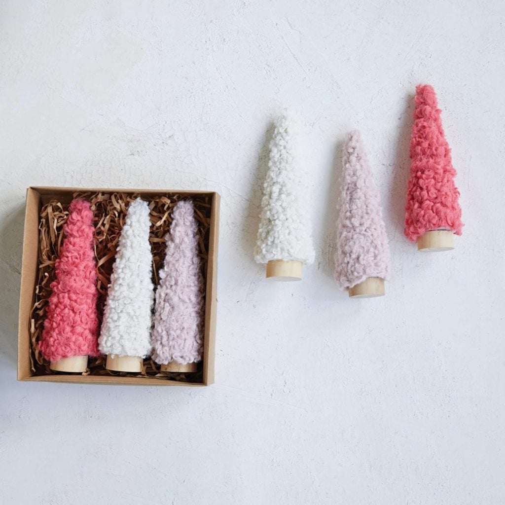 Four small cone-shaped decorative trees with textured surfaces in pink, white, and lavender evoke a whimsical pink Christmas vibe, shown both inside and outside a cardboard box with packing material.