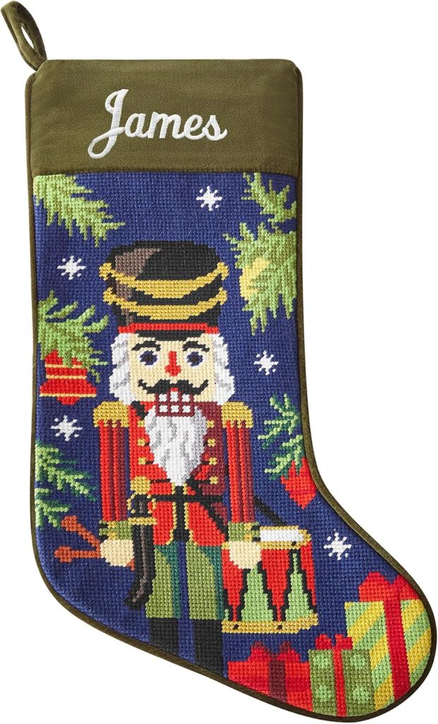 A Christmas stocking with "James" embroidered on top, featuring a needlepoint nutcracker, drum, gifts, and ornaments on a blue background—perfect for bringing Christmas Morning magic to your home.