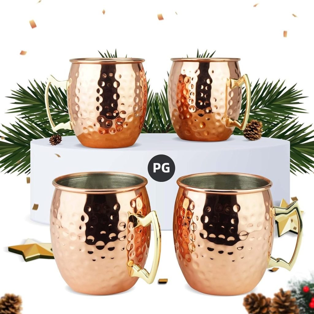 Four copper mugs with hammered texture on display, surrounded by pine branches and scattered pinecones.