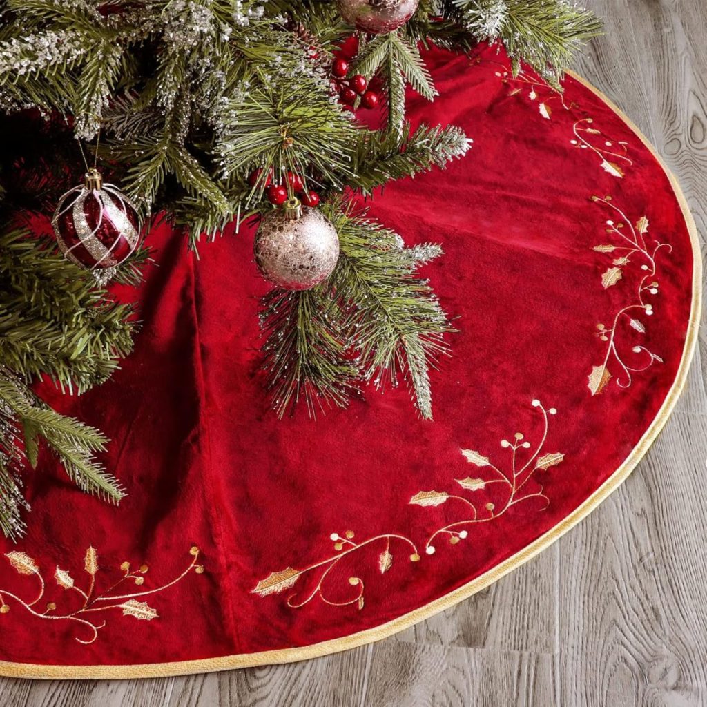 Close-up of a Christmas tree adorned with red and gold ornaments. It stands elegantly on a red and gold embroidered Christmas tree skirt, perfectly complementing the wooden floor beneath it.